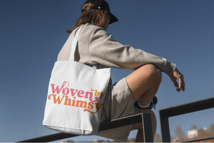 Woman sitting on railing with white canvas bag over her shoulder with "Woven By Whimsy" printed in pink ink.